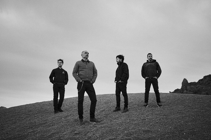 Four men stand apart on a low, rocky hill under an overcast sky, mid-stance and observant of the distance.