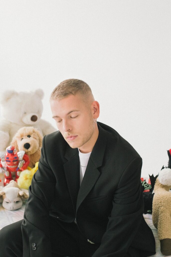 Young man in a black blazer seated among plush toys, eyes closed, calm pose in a bright room