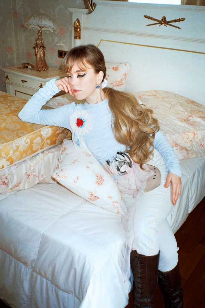 Young woman with long blonde hair sits on a bed in a vintage pastel bedroom, wearing a light blue top and brown knee-high boots.