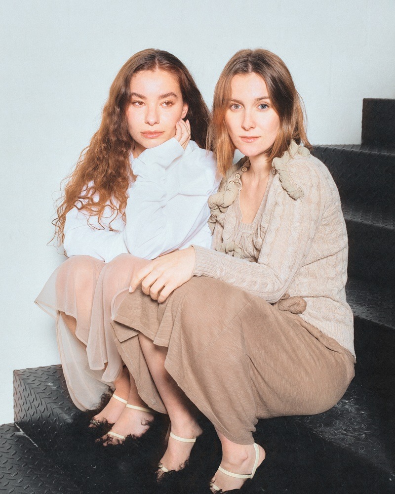 Two women sit close together on dark stairs, looking at the camera in soft beige and cream outfits.
