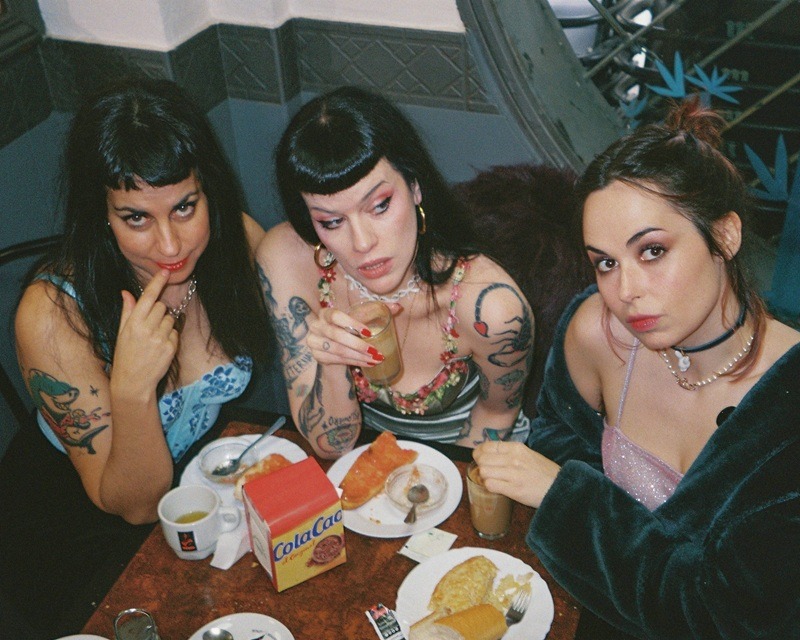 Three tattooed women sit at a table with drinks and plates of food, posing for the camera.
