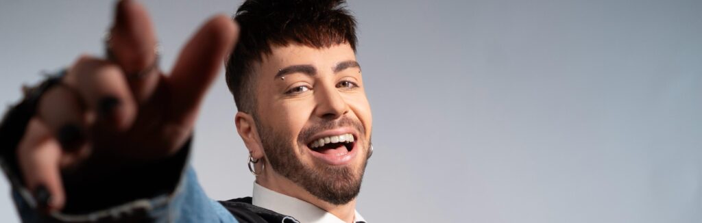 Close-up portrait of a smiling man with a beard and facial piercings, reaching toward the camera.