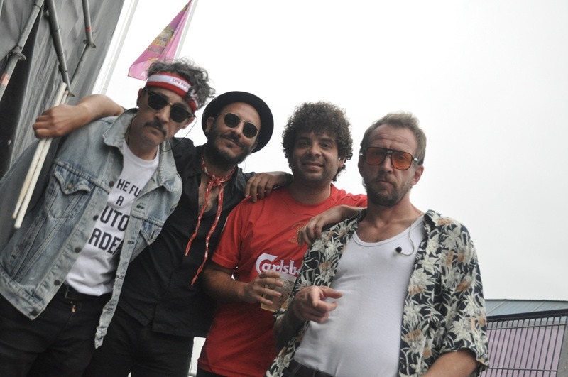 Four men stand shoulder to shoulder at what looks like a festival, posing for the camera. From left: a man in a denim jacket and headband with sunglasses; a man in a black hat and beard; a man in a red Carlsberg T-shirt holding a cup; and a man in a tropical-pattern shirt with orange-tinted glasses.