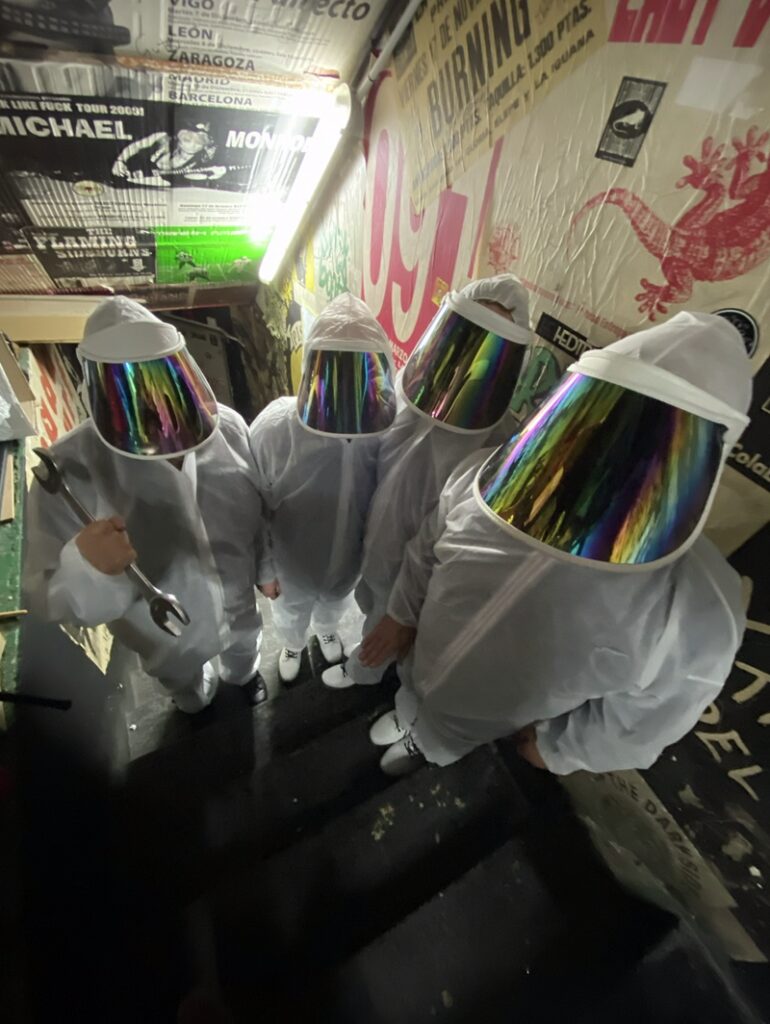 Four people in white hazmat suits with rainbow reflective visors stand on a stairwell covered in posters and graffiti.
