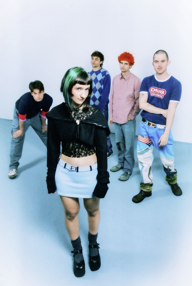 Frontwoman with green-tinted hair, black lace top and light blue skirt, standing confidently in front of four male band members in a studio.