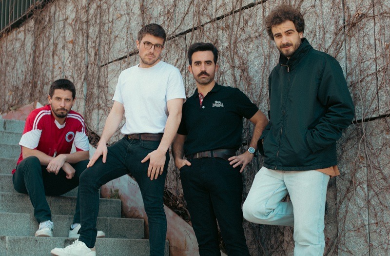 Four men pose on concrete stairs outdoors against a wall covered in vines, casually dressed and looking at the camera.