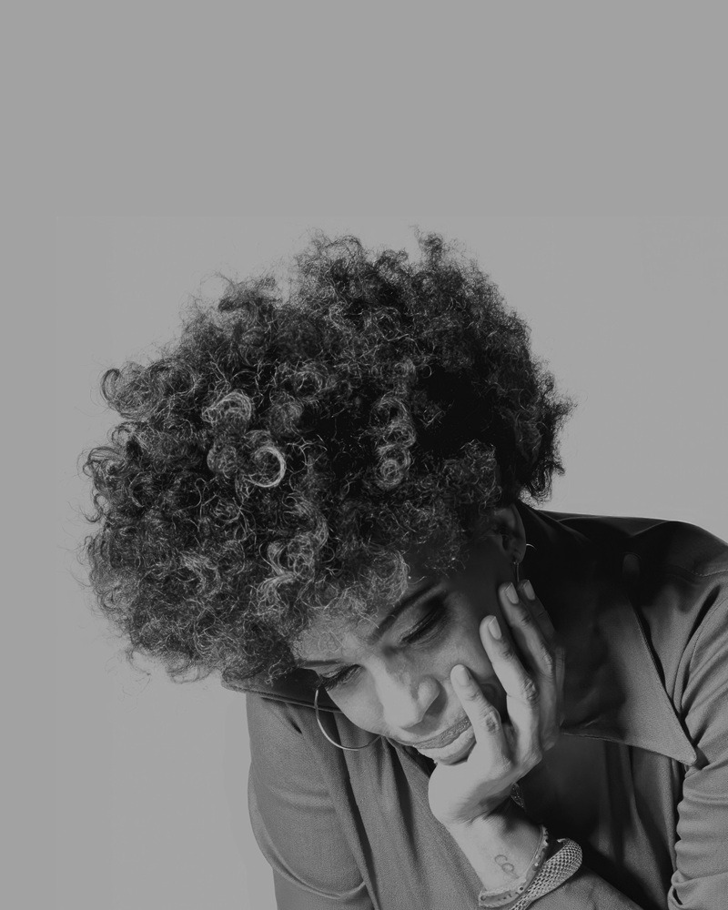 Woman with curly hair resting her head on her hand, looking down thoughtfully (black-and-white portrait). Alternate: Person with voluminous curls leaning on hand in a contemplative pose.