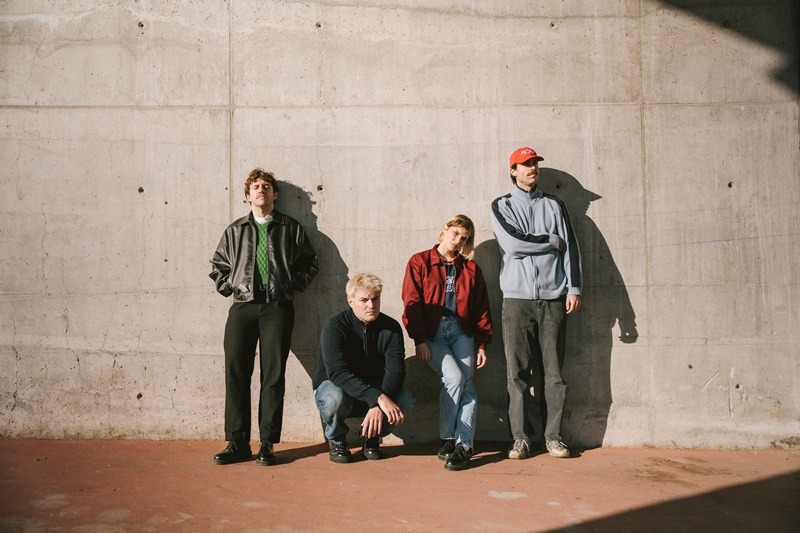 Four men in casual jackets posing against a concrete wall; one crouches while the others stand with relaxed poses in daylight.