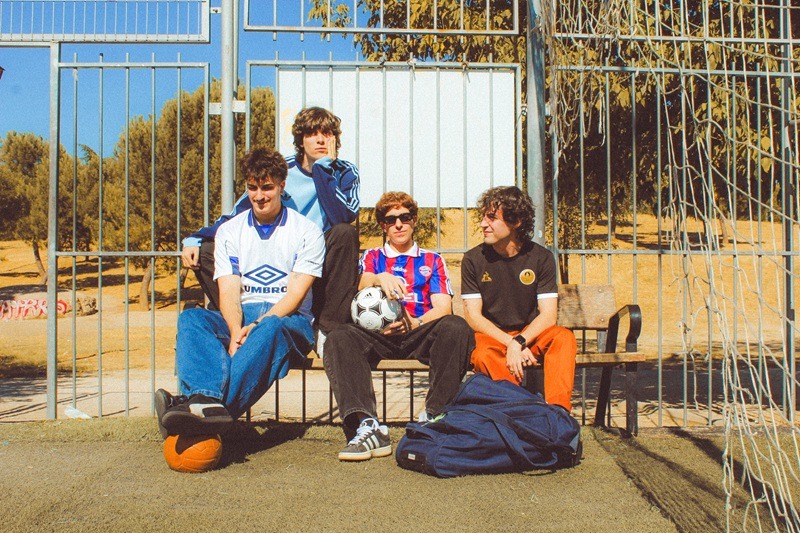 Five young men sit on a bench by a metal fence on a sunny outdoor sports area, with a soccer ball at their feet and a backpack nearby.