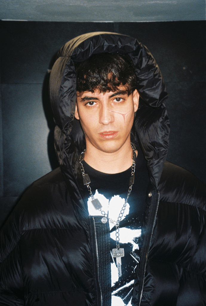 Young man in a black hooded puffer jacket with a silver chain necklace, facing the camera; visible facial scar on his left cheek.