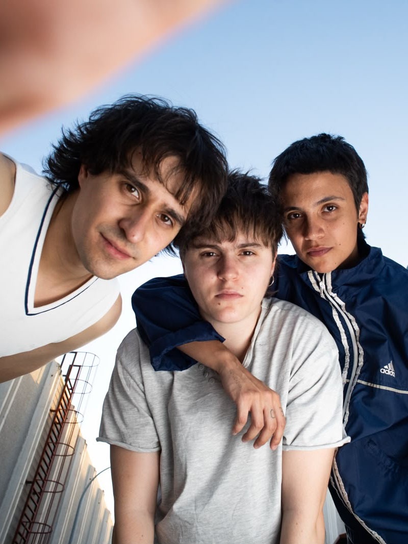 Three friends posing for a selfie outdoors against a clear blue sky; one has an arm around a shorter friend in a gray shirt in the center.