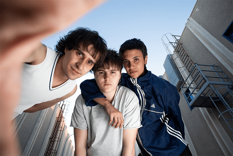 Three young men pose for a low-angle selfie, standing close together in an urban stairwell area.