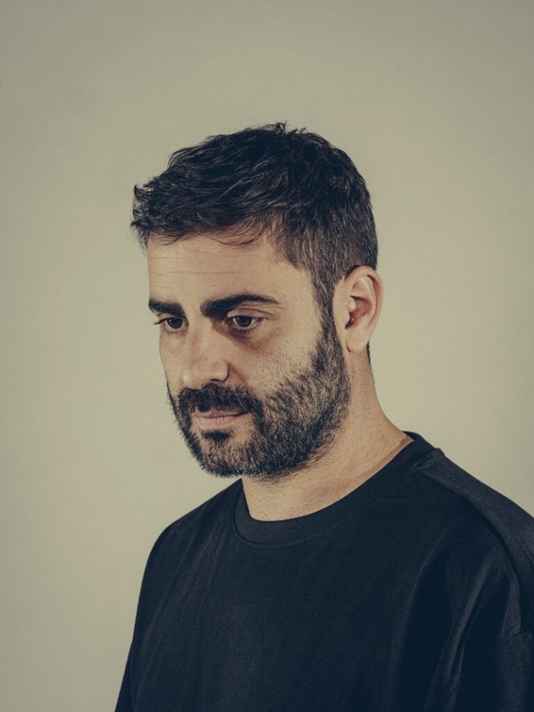 Portrait of a man with dark hair and a full beard, wearing a black crew-neck shirt, looking down in a contemplative pose.