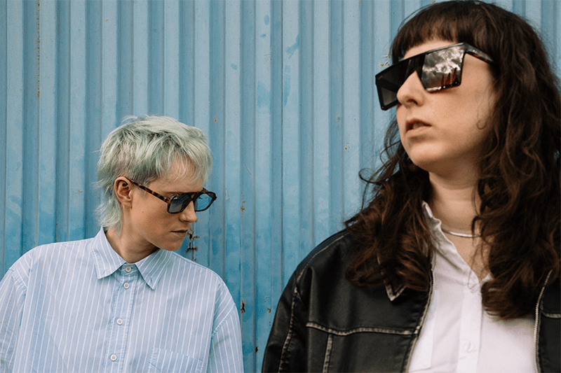 Two people wearing bold sunglasses stand against a blue corrugated metal wall; one with short light hair in a striped shirt, the other with long dark hair in a leather jacket.