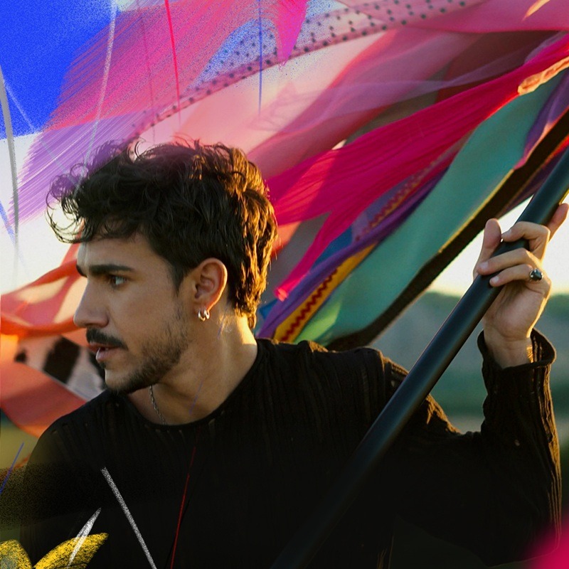 Young man with dark hair and stubble holding a black pole as vibrant fabric banners flutter around him.",