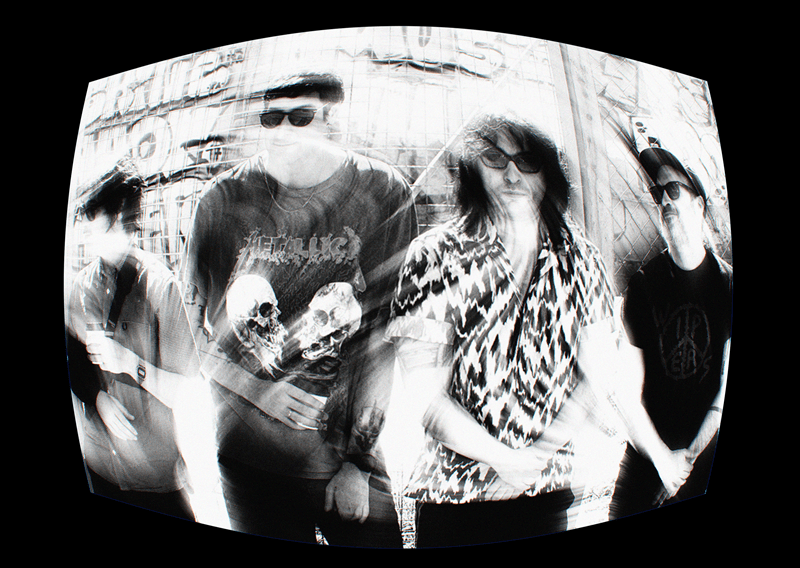 Black-and-white distorted photo of four rock musicians wearing sunglasses, posing against a graffiti-covered wall with a Metallica shirt visible on one member